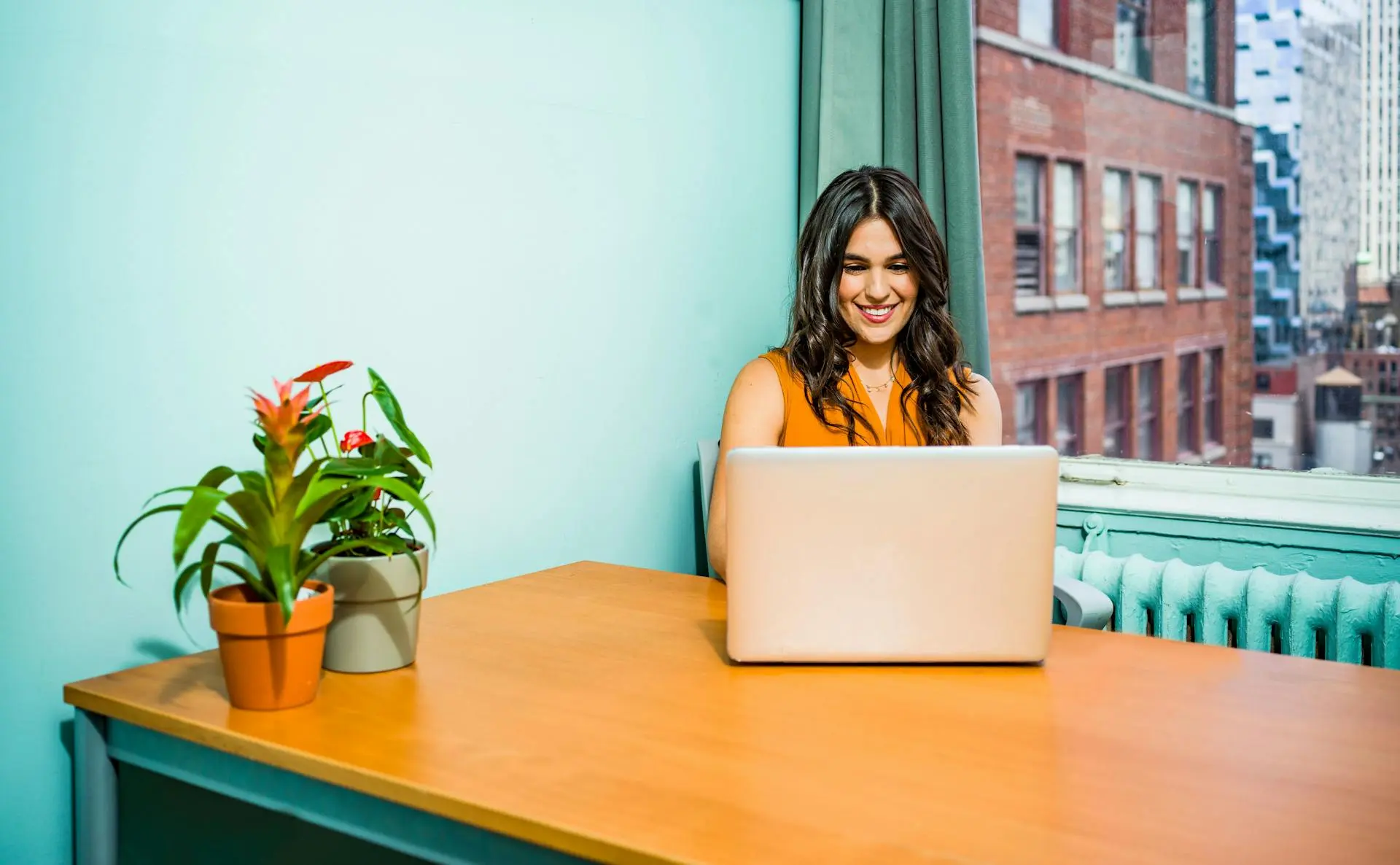 A woman in an orange top sits at a desk with a laptop searching "What to Sell with Print-on-Demand", smiling. Plants in pots are on the desk. A window shows a cityscape outside. Bright, cheerful setting.