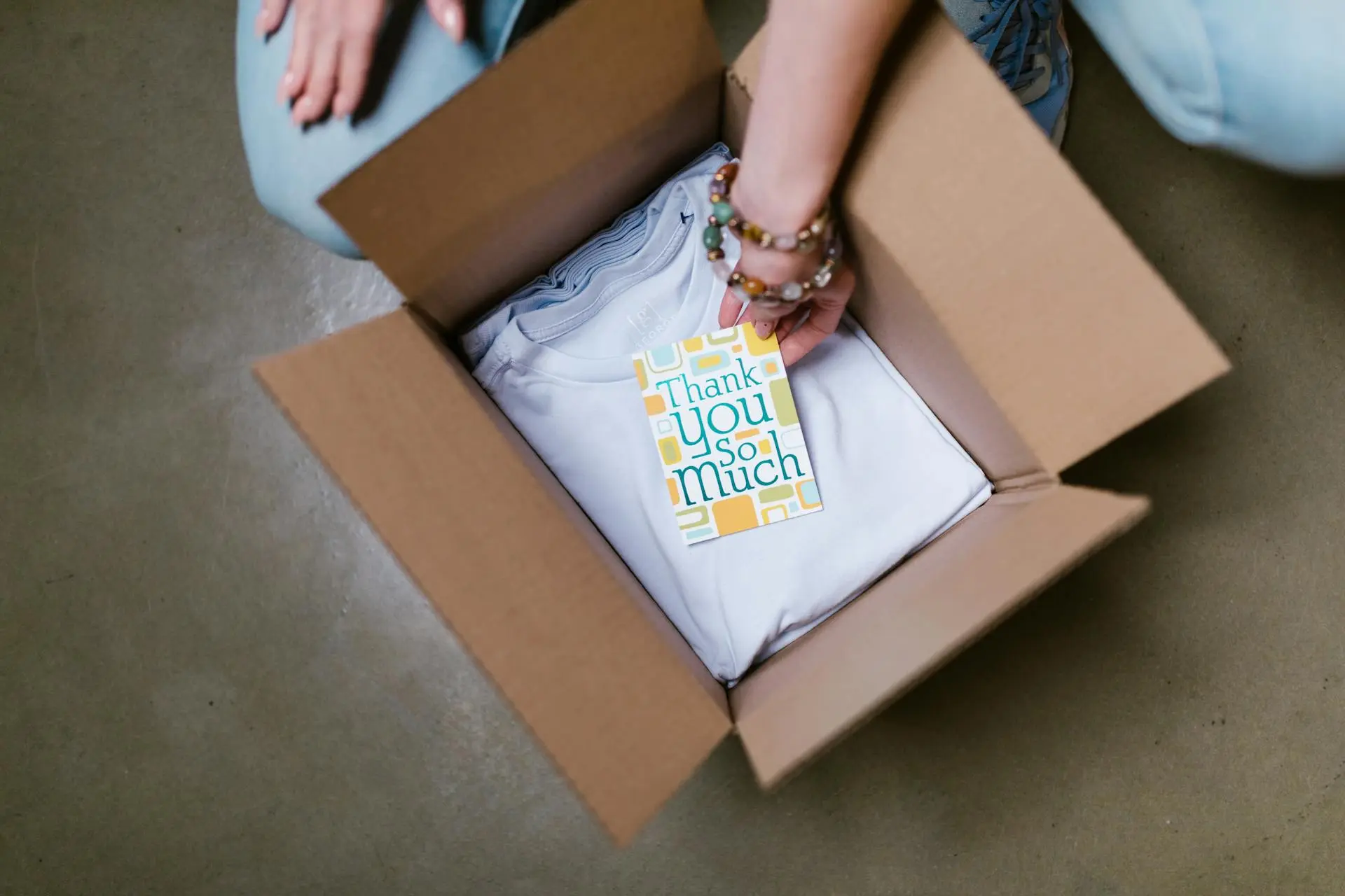 A person places a colorful "Thank You So Much" card on a neatly folded white shirt inside an open cardboard box, on a gray surface.